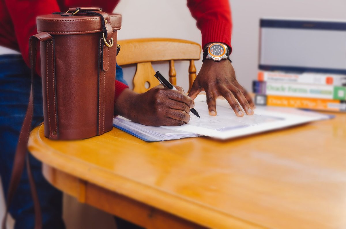 Une personne debout devant une table écrit au stylo sur un document posé à plat, un sac et une pile de livres à côté.